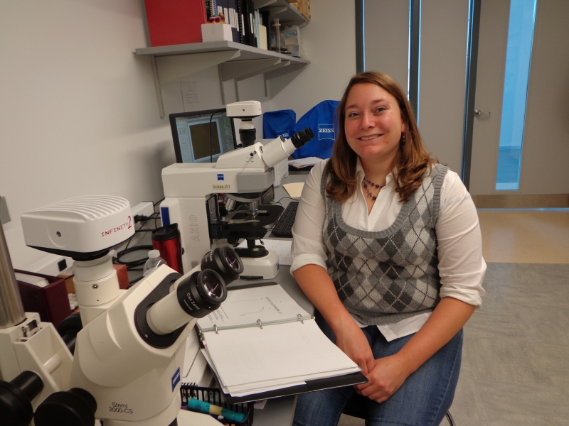 A smiling person sits near a counter with two microscopes in a lab. A binder is open in front of her.