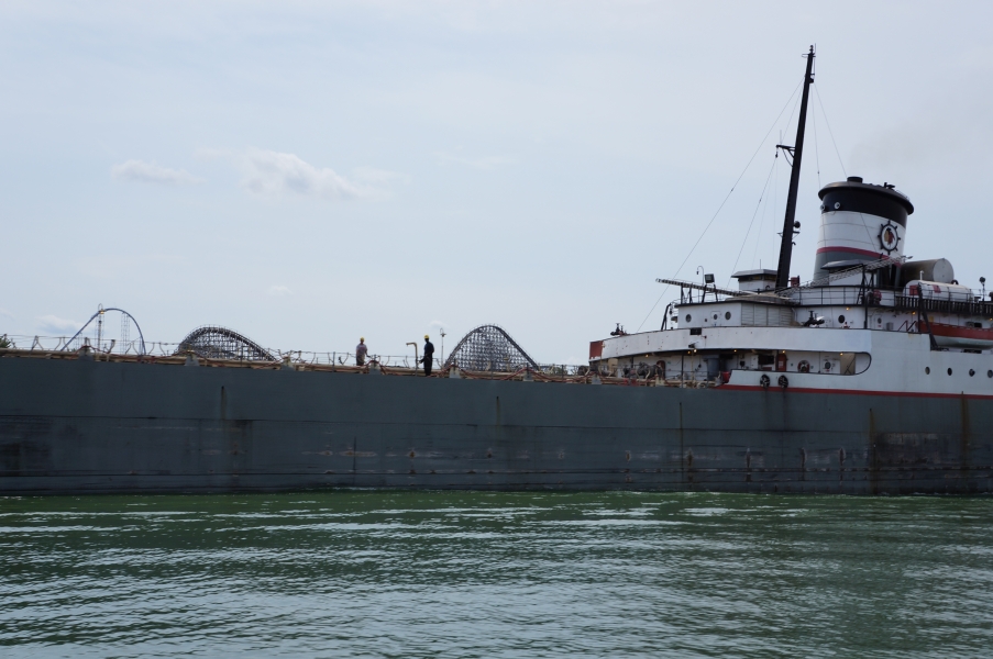 People working on the flat deck of a long large boat. There is a few decks at the back and a smoke stack.