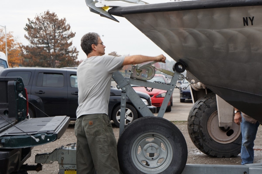 A person stands by a boat on a trailer turning a crank to tighten a strap between the trailer and the boat.