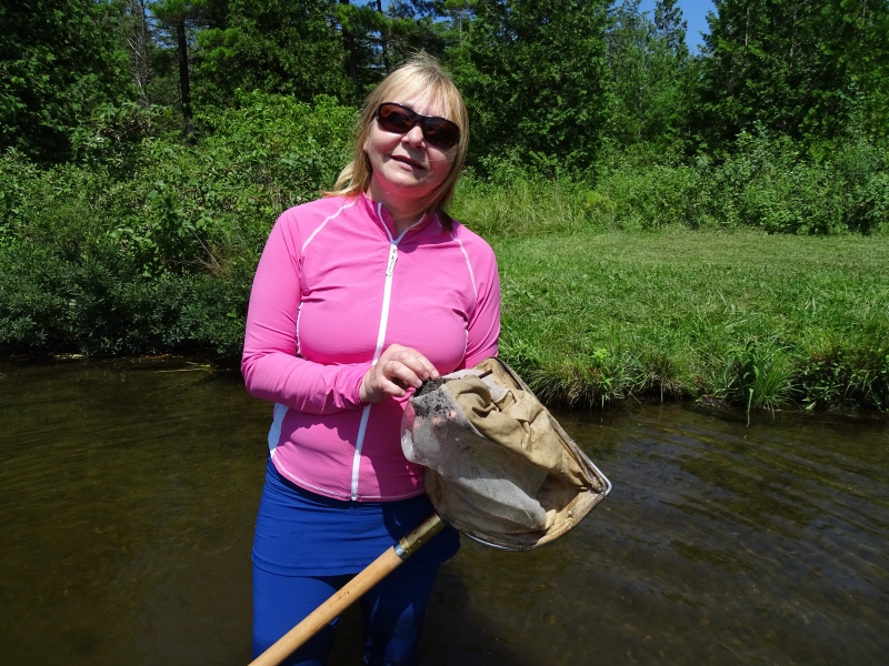 A person wearing sunglasses stands in a creek holding a net in her hands.