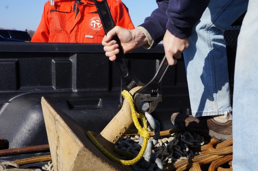 Someone uses two large wrenches to secure a shackle between a pyramid anchor and a heavy chain lying in the bed of a truck