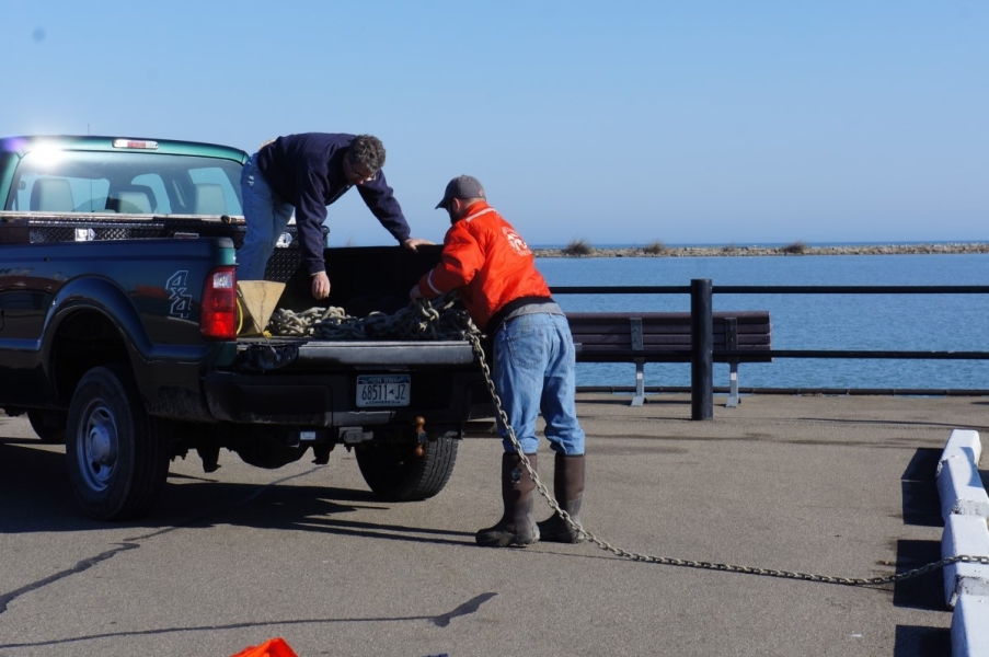 Two people pulling a chain off the back of a pickup truck. They are near water.