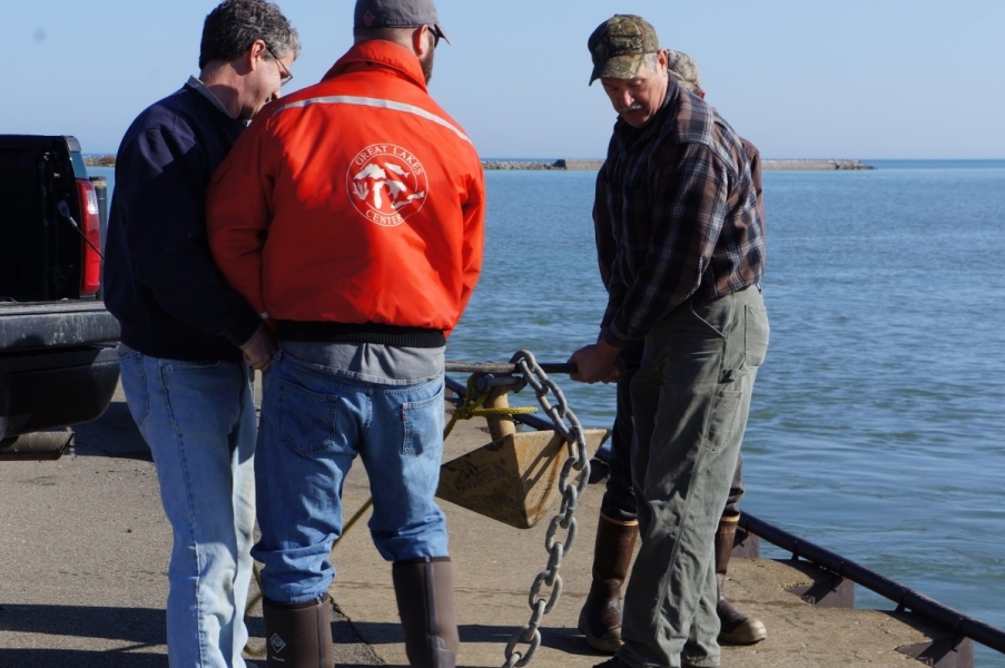 Four people lift a pyramid anchor on a metal rod. They are near water.