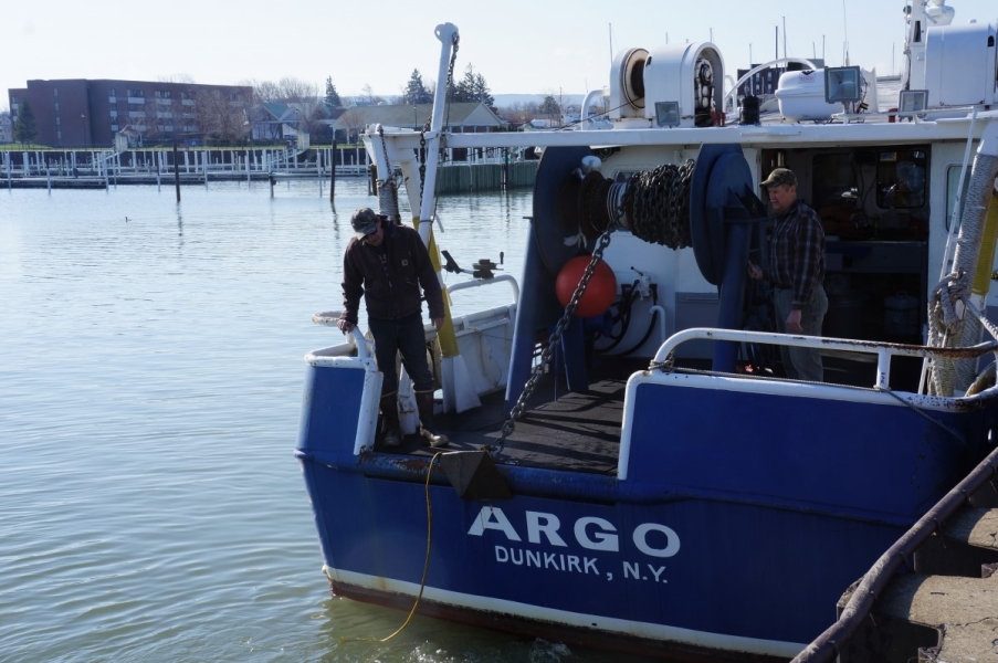 Two people coil chain onto a large reel on the back of a boat. The anchor is just below the deck. The boat is named "Argo"