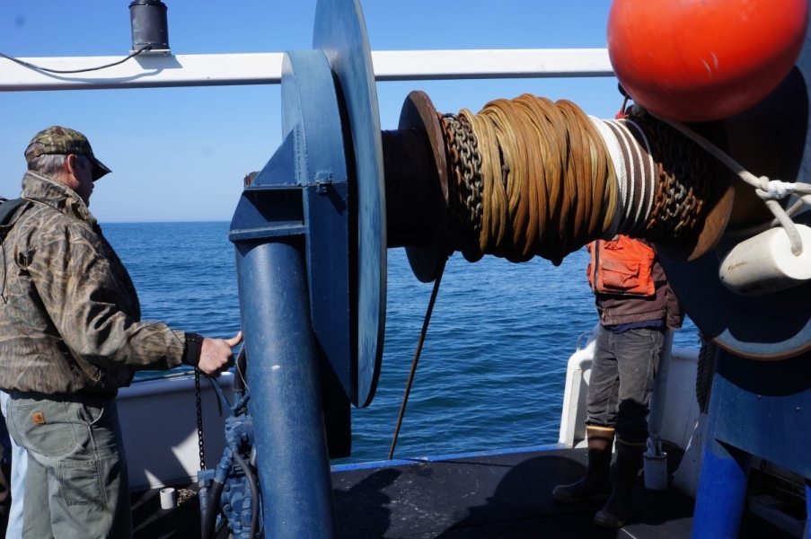 A person operates a large reel on a boat to lower a rope into the water