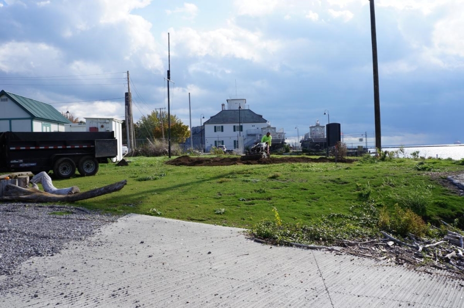 A person pushes a tilling machine in an area of dirt in a lawn near the waterfront by a telephone pole and a boat ramp.