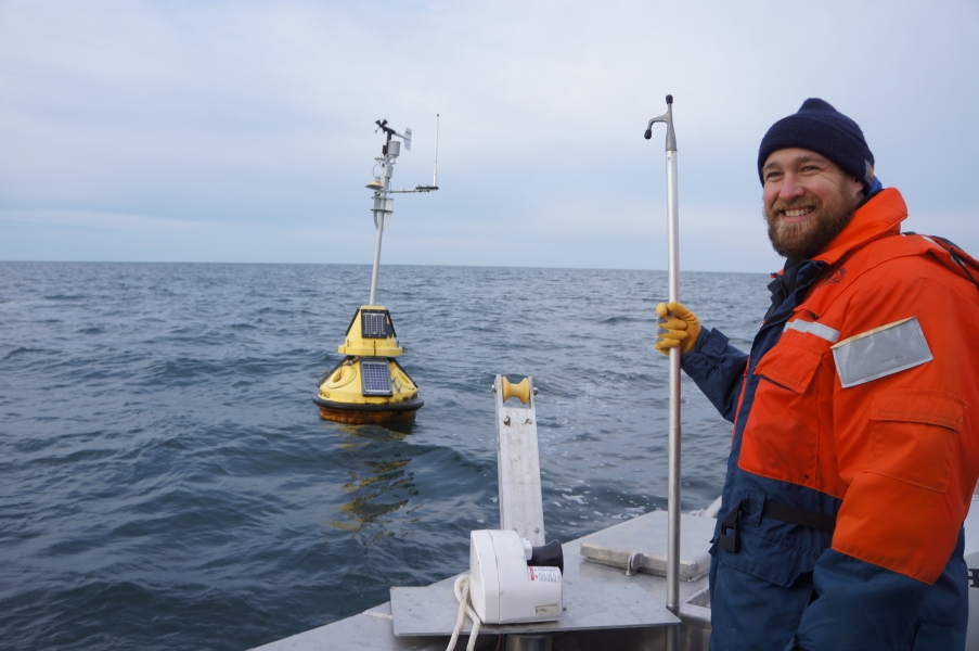 A smiling person in winter gear stands on the back deck of a boat, holding a boat hook. There is a buoy in the water nearby and a pot puller winch on the side of the boat.