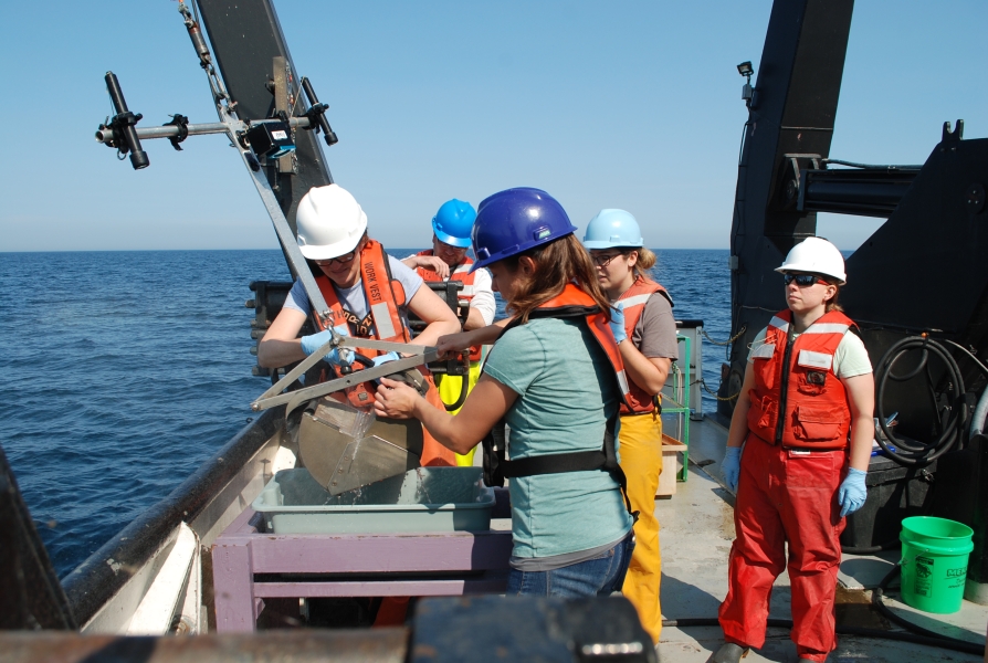 Two people open up a grab sampler into a bucket on the deck of a large boat. Three people are standing behind them.