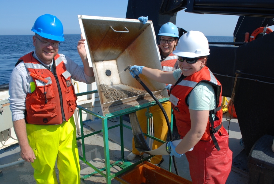 Two people tilt up a basin with sediment in it while a third person uses a hose to wash the sediment through a hole into a mesh sleeve with a bottle on the end of it. They are working on the deck of a large boat.