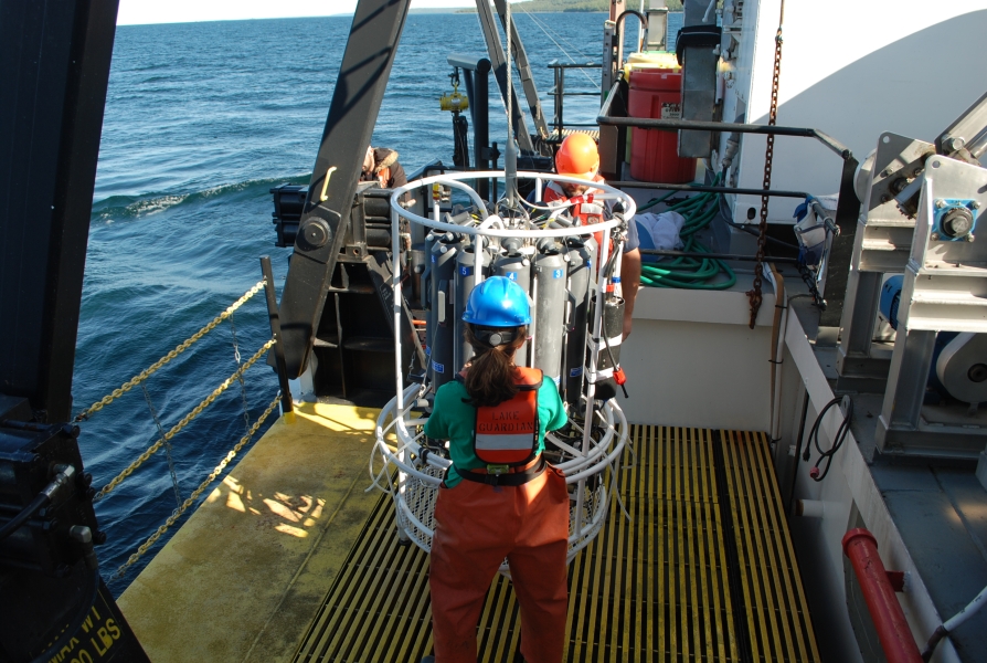 Two people stand on the deck of a large boat to prepare a large piece of equipment to be deployed.