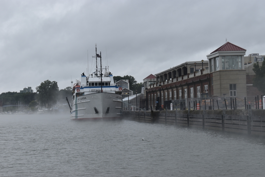 The bow of a large boat that is docked in a misty river by a commercial building