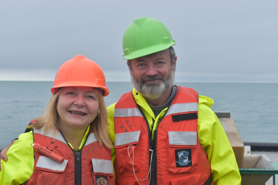 Two people pose for a picture while on the water on a cloudy day. They are both wearing safety gear: hard hats, life jackets, and rain coats.