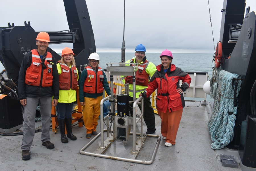 Five people stand around a piece of equipment in a metal frame on the back of a large boat.