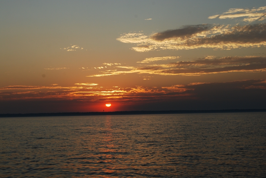 A sunset over water. A bright red sun peaks through a few white clouds.