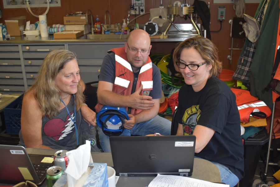 Three people sit in front of a laptop in a lab on a boat. One person is wearing a life jacket.