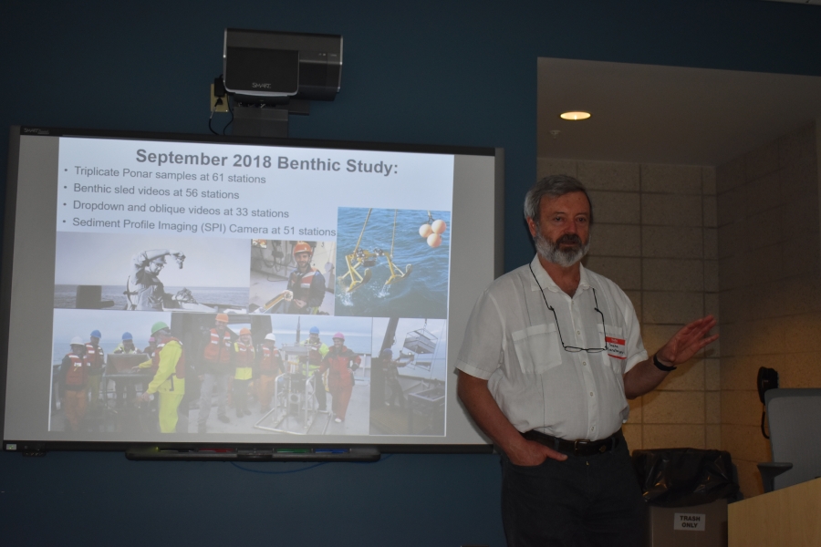 A person stands in front of a projector screen giving a talk. The slide on the screen has pictures and says "September 2018 Benthic Study."