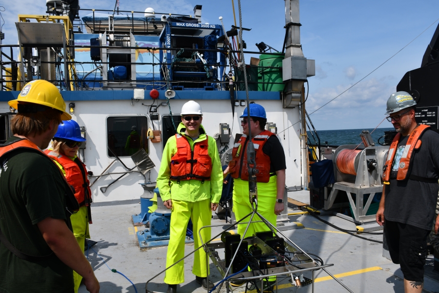 Several people in hard hats and life jackets stand around a piece of equipment on the deck of a large boat