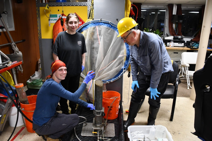 Three people gather around a sediment core inside a room on a boat, two smiling while the other looks at the core. One person is kneeling next to the core and measuring it with a tape measure