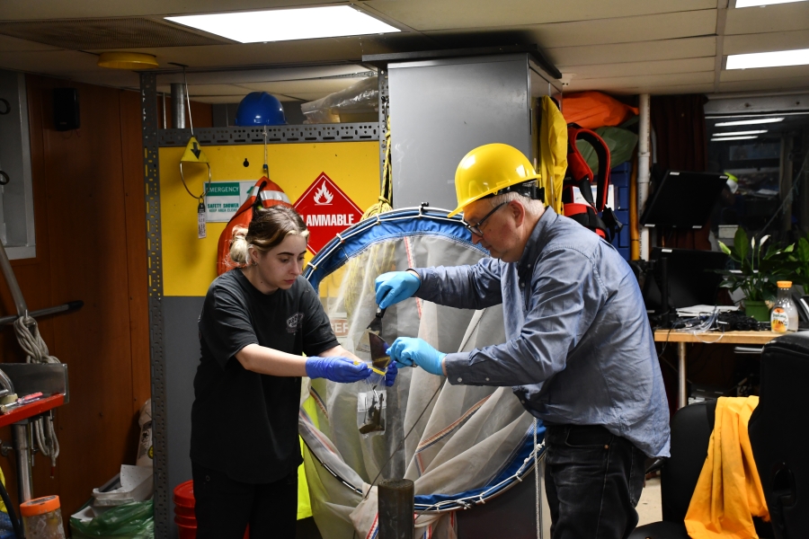 Two people work to collect a sediment sample from a sediment core in a room on a boat. One person holds the sample bag while a second person scrapes mud from putty knives into the bag