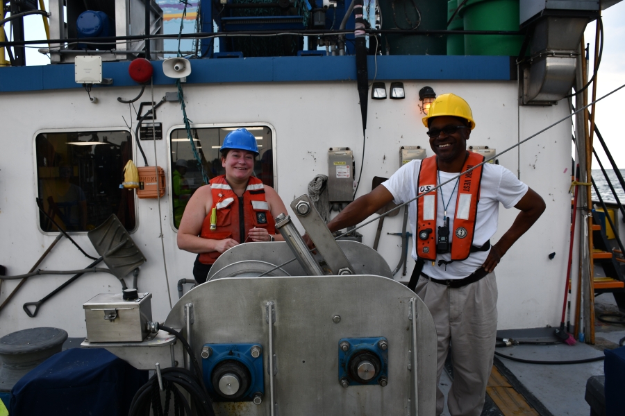 Two people in life jackets and hard hats smile and stand next to a winch on the deck of a large boat