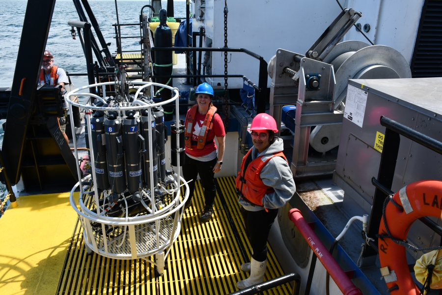 Three people in life jackets and hard hats smile and work on the deck of a boat by a rosette of water sampling bottles