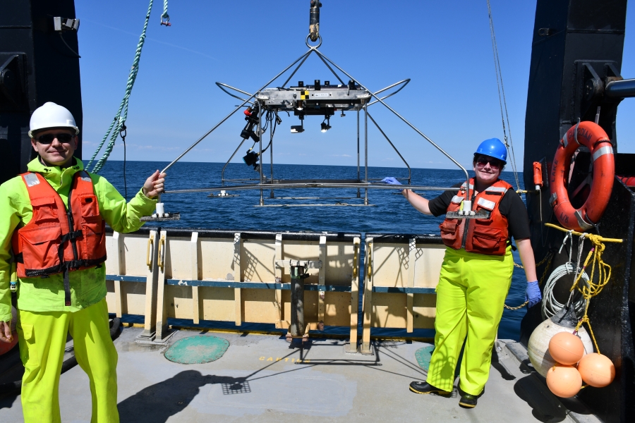 Two people in life jackets and hard hats smile and hold a large frame with equipment as it's being held up by a winch on a large boat