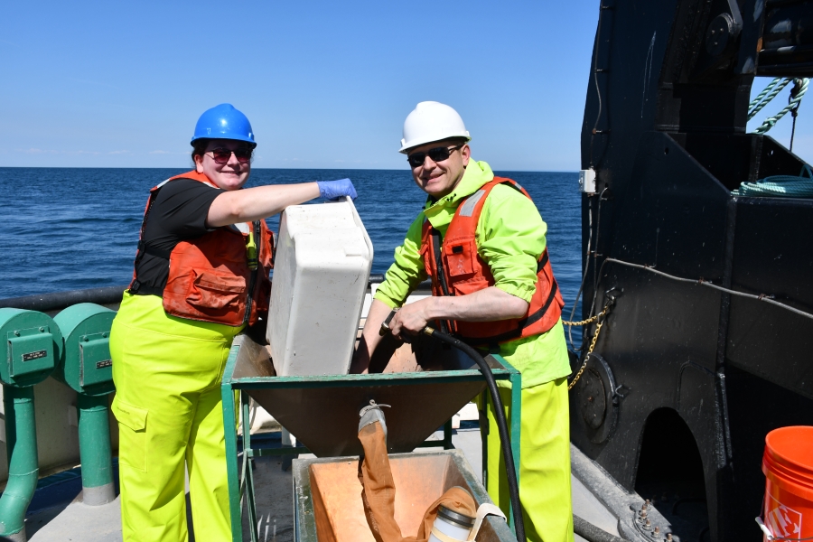 Two smiling people in life jackets and hard hats work at a basin on the deck of a large boat. One person dumps a tub into the basin while the other uses a hose to wash the tub