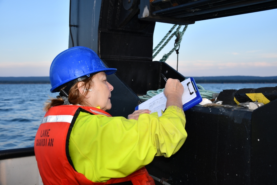 A person in a life jacket and hard hat writes on a clipboard braced on a large piece of hydraulic equipment on a large boat