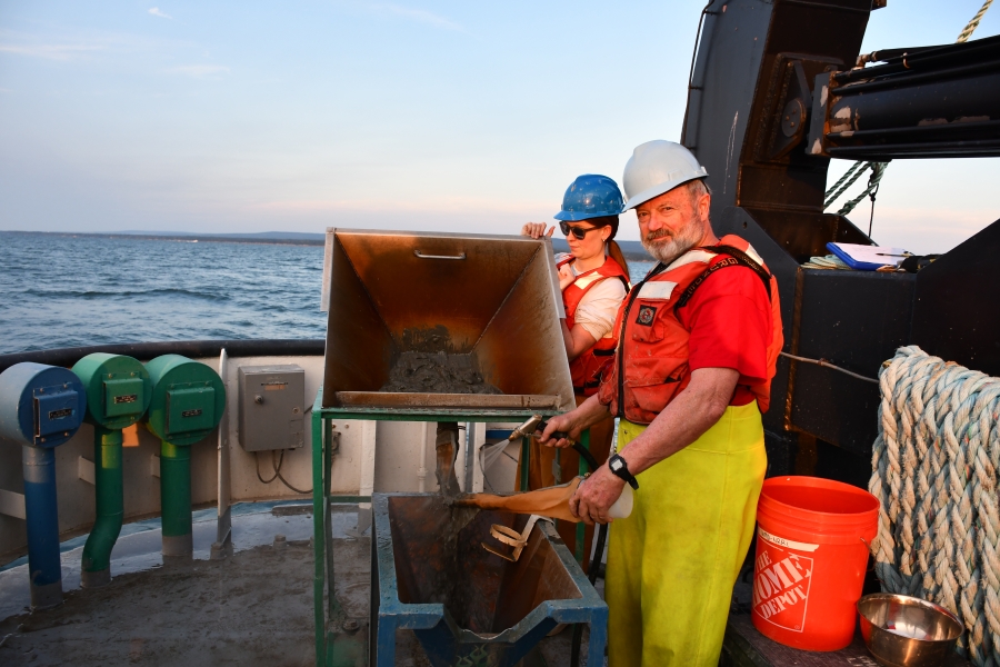 Two people in life jackets and hard hats work at a basin on the deck of a large boat at sunset. One person is lifting the basin so the muddy water runs into a mesh tube that the other person is washing with a hose to remove fine sediment, with a sample jar at the bottom of the mesh tube.