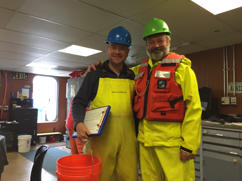 Two people wearing safety gear pause pose for a picture inside a lab on a boat. They are wearing rain gear and hard hats. One has a life jacket, and the other has a clipboard and a bucket.