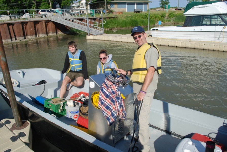 A person stands at the console of a small motor boat tied to a dock in a marina. Two other people sit on the rail. There is gear in the boat.