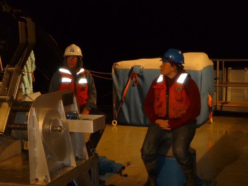 Two people in hard hats and life jackets sit on the back deck of a boat at night