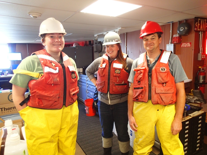 Three people in a lab on a boat wearing hard hats, life jackets, and rain pants.