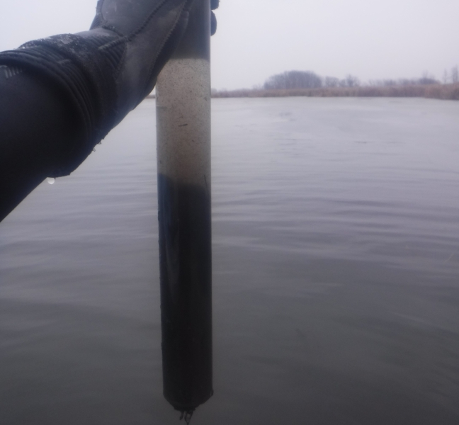 A gloved hand holding up a tube of water and sediment above a body of water on an overcast day