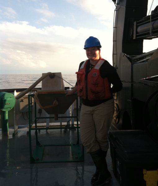 A person in a hard hat and life jacket stands on the back deck of a boat next to a metal table with something on it.